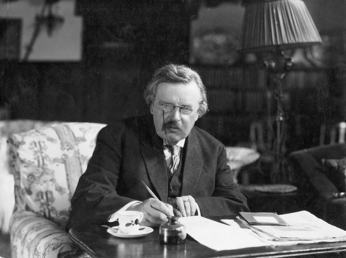 G.K. Chesterton seated at a writing table with a pen in hand, a teacup beside him, in a book-lined room.