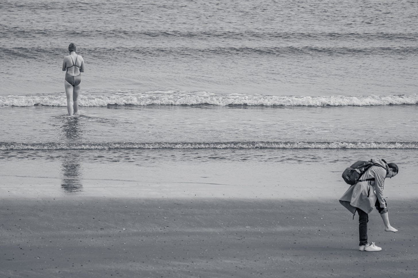 A lone figure walking out into the shallows of a wide flat sea, in black and white, low waves running across the frame. A lone figure walking out into the shallows of a wide flat sea, in black and white, low waves running across the frame.