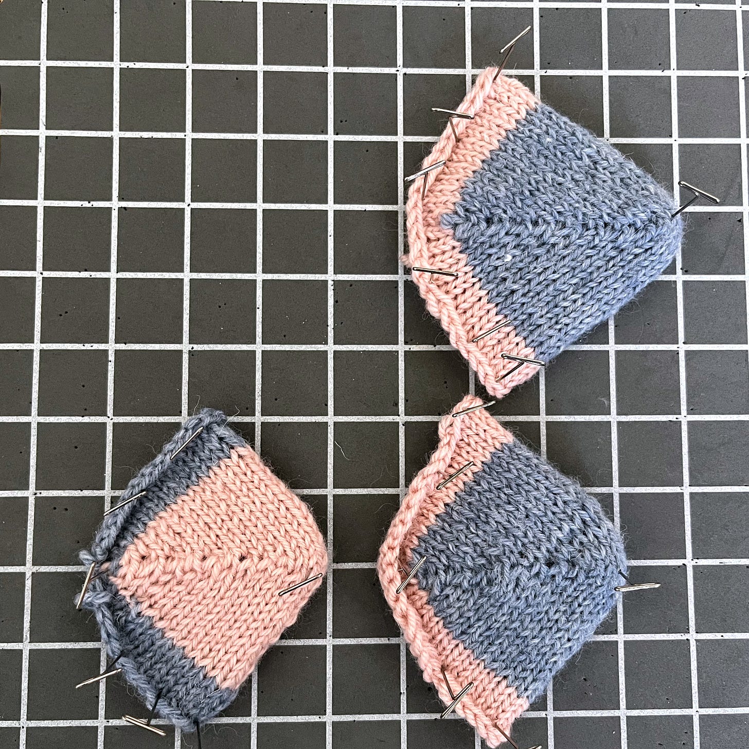 Four small knitted swatches in blue-grey and pink yarn, pinned to a grey foam blocking mat. Top row: two standard short row toes. Bottom row: two rounded short row toes. Left column worked cuff-down; right column toe-up. Four small knitted swatches in blue-grey and pink yarn, pinned to a grey foam blocking mat. Top row: two standard short row toes. Bottom row: two rounded short row toes. Left column worked cuff-down; right column toe-up.