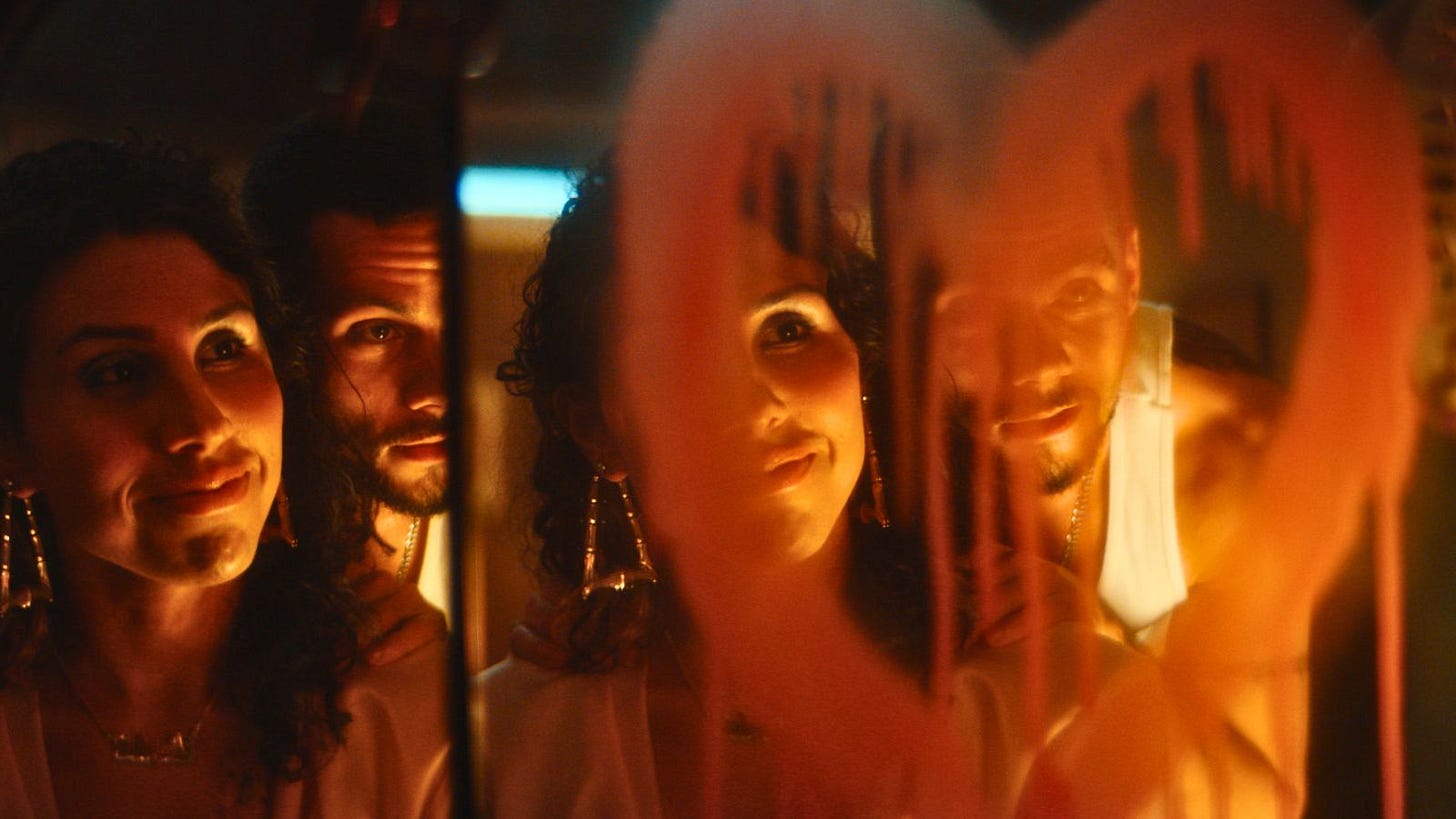 Two people look into a trifold mirror. Dried red spray paint in the shape of a heart distorts the two figures in the middle mirror. The room is dimly lit by cold fluorescent lights and warm candlelight. Movie still from Ponyboi
