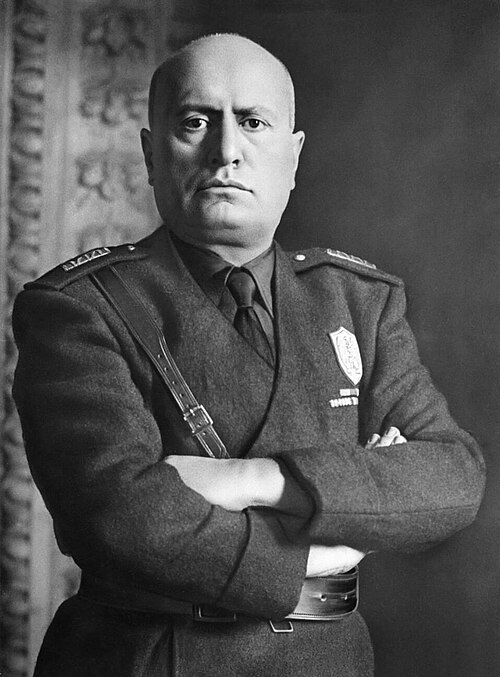 Benito Mussolini in a military uniform with a shield-shaped badge on the chest, arms crossed, wearing a dark jacket with epaulets and a tie, standing against a backdrop with ornate patterns. Text on the badge reads "Milizia Volontaria per la Sicurezza Nazionale".