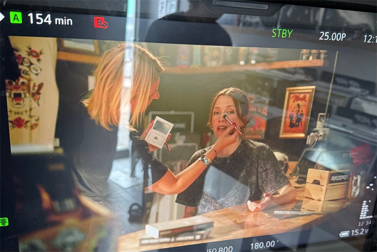The preview monitor on a studio set shows a blonde woman touching up the makeup of the presenter on screen who wears a grey t-shirt and has tied back brown hair. The preview monitor on a studio set shows a blonde woman touching up the makeup of the presenter on screen who wears a grey t-shirt and has tied back brown hair.