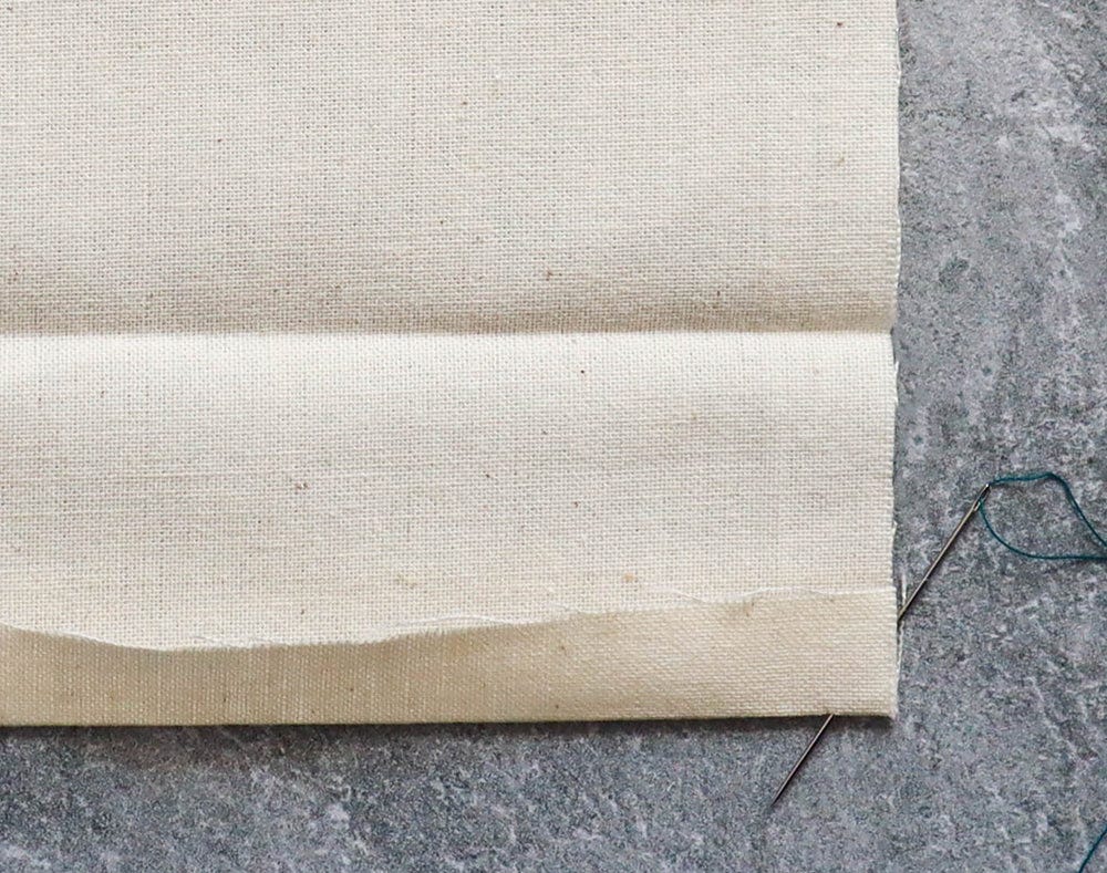 Cream muslin fabric with folded hem and needle threaded with teal thread