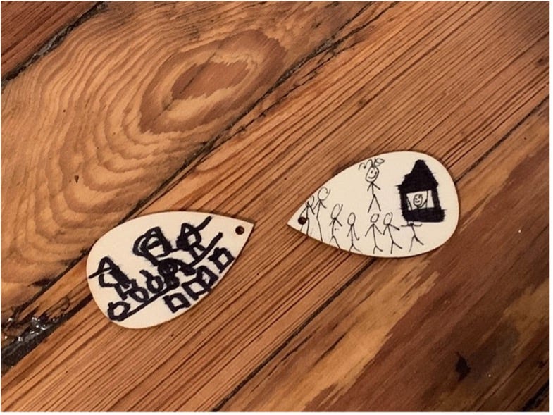 Two teardrop shaped wooden pendants with original artwork. On the left, a table of pumpkins and books. On the right, a canopy tent and stick figures in a line, one of them labeled "Me."