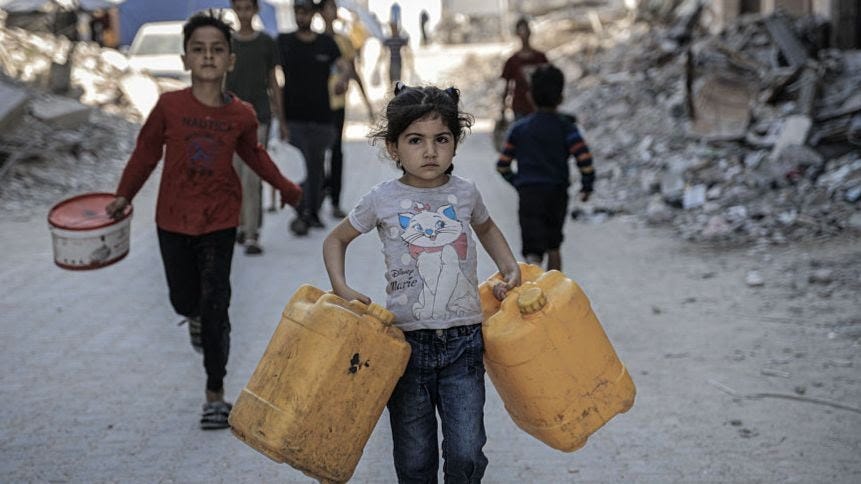 Children walk along a road carrying containers. Large piles of rubble can be seen in the background. A young girl wears a T-shirt with a cat on it and jeans, a boy wears black trousers and a red top