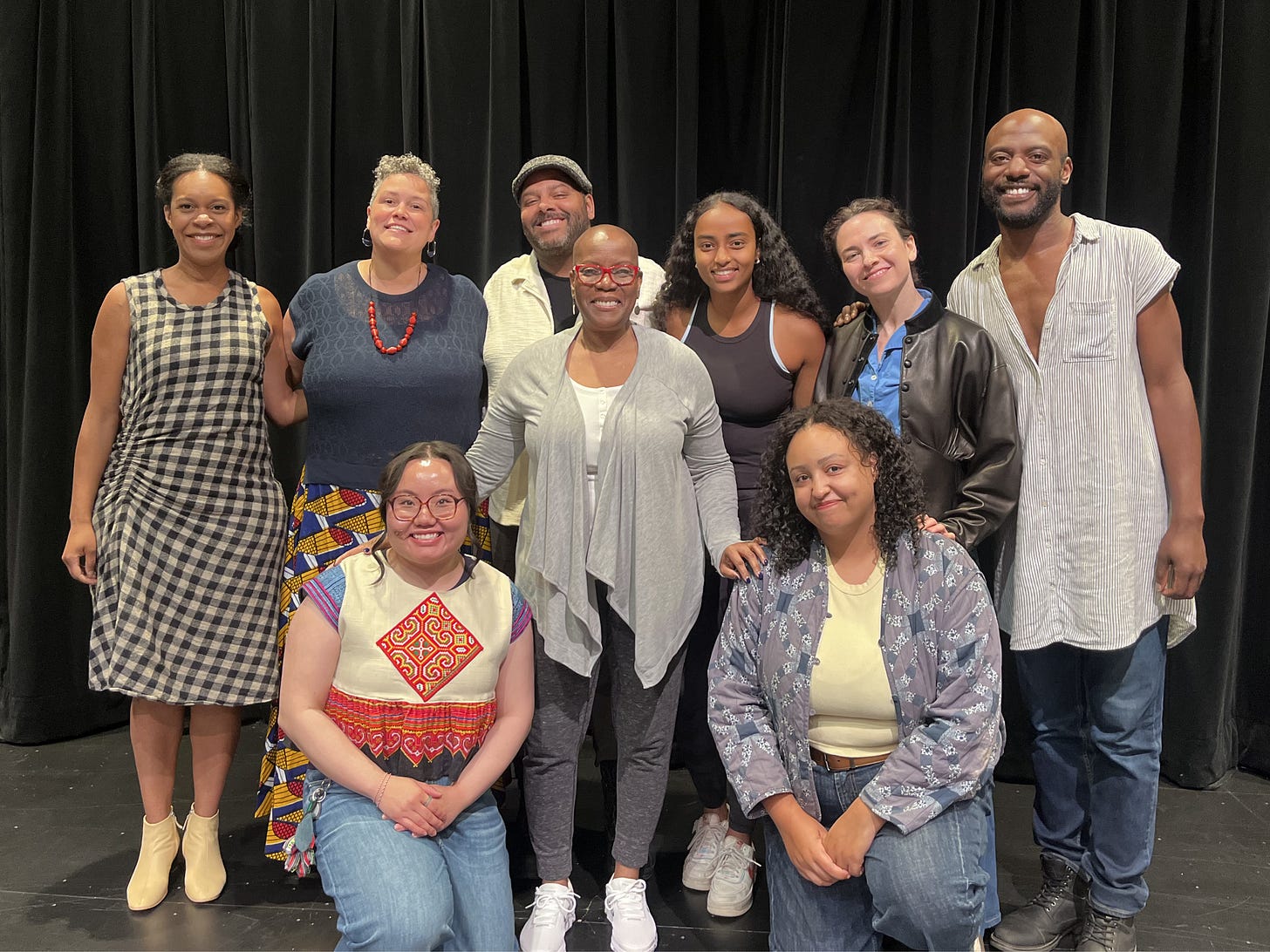Group photo of the creative team behind The Quilted Heart following a workshop at Playwrights' Center. Nine smiling artists pose together on a black-box theatre stage in casual rehearsal clothes, standing and kneeling in two rows in front of black curtains. The atmosphere is warm, celebratory, and collaborative.