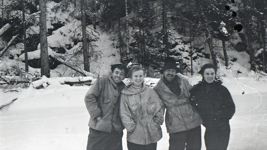 An old black and white photo of four young people standing in front of a row of pines. They are smiling and wearing winter clothing and there is snow all around them.