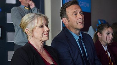 A man and woman sit at a school assembly smiling
