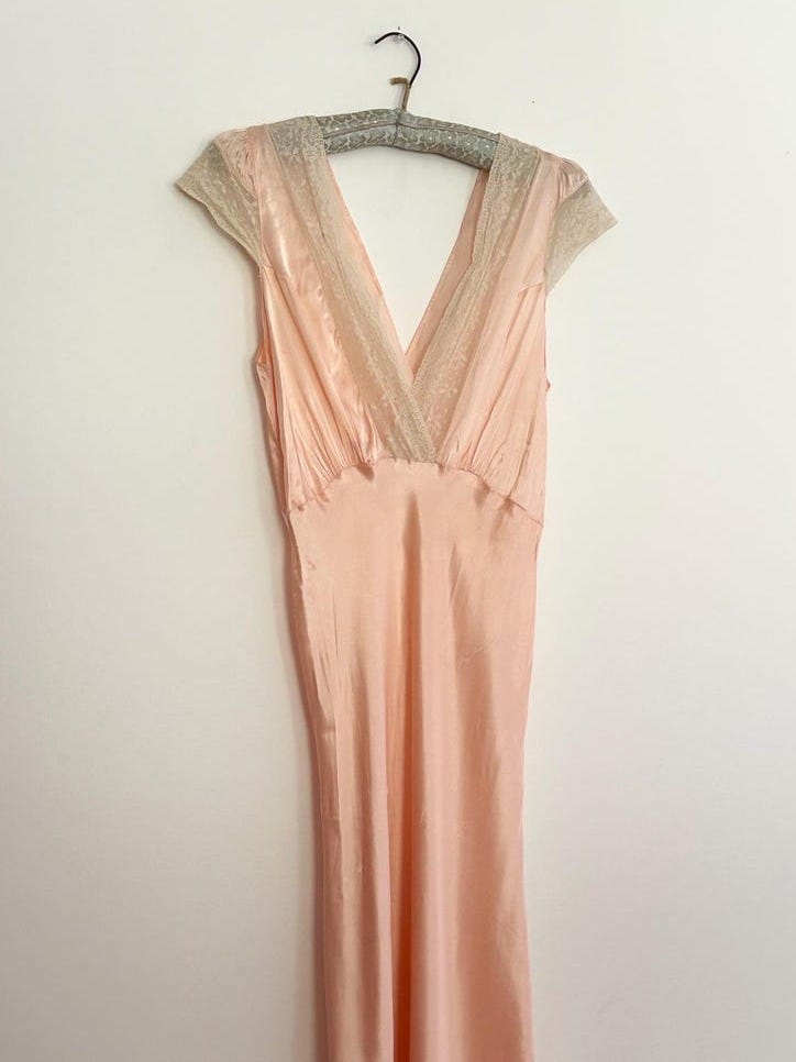 May include: A floor-length pale peach satin dress featuring a deep V-neckline trimmed with delicate cream-colored lace. The garment has short cap sleeves and a smooth, flowing silhouette that hangs straight down from the bodice to the hemline.