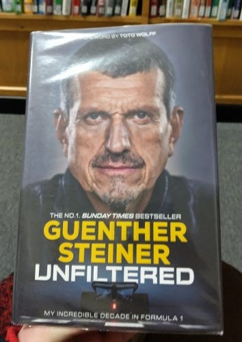 A photo of a hardback edition of 'Unfiltered.' There's a row of library books in the background and grey library carpet, too.