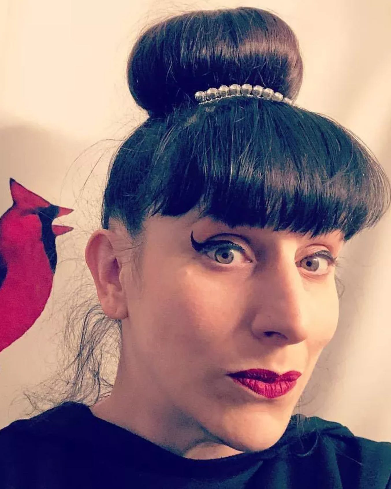 Headshot of Kat Kinsman, a white woman with dark hair swept up in a high bun and deep red lipstick. A cardinal (painted on the wall behind her?) peeks into frame.