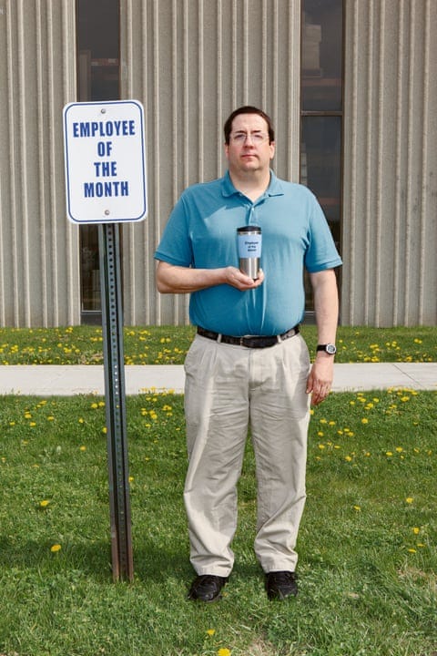 A man standing next to an Employee of the Month sign, holding a trophy in his hand