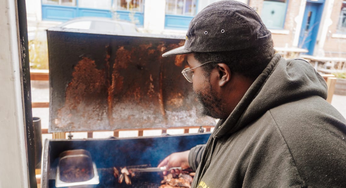 A man - Jowayne Marks - wearing a black hoodie and manning a barbecue with meat on it
