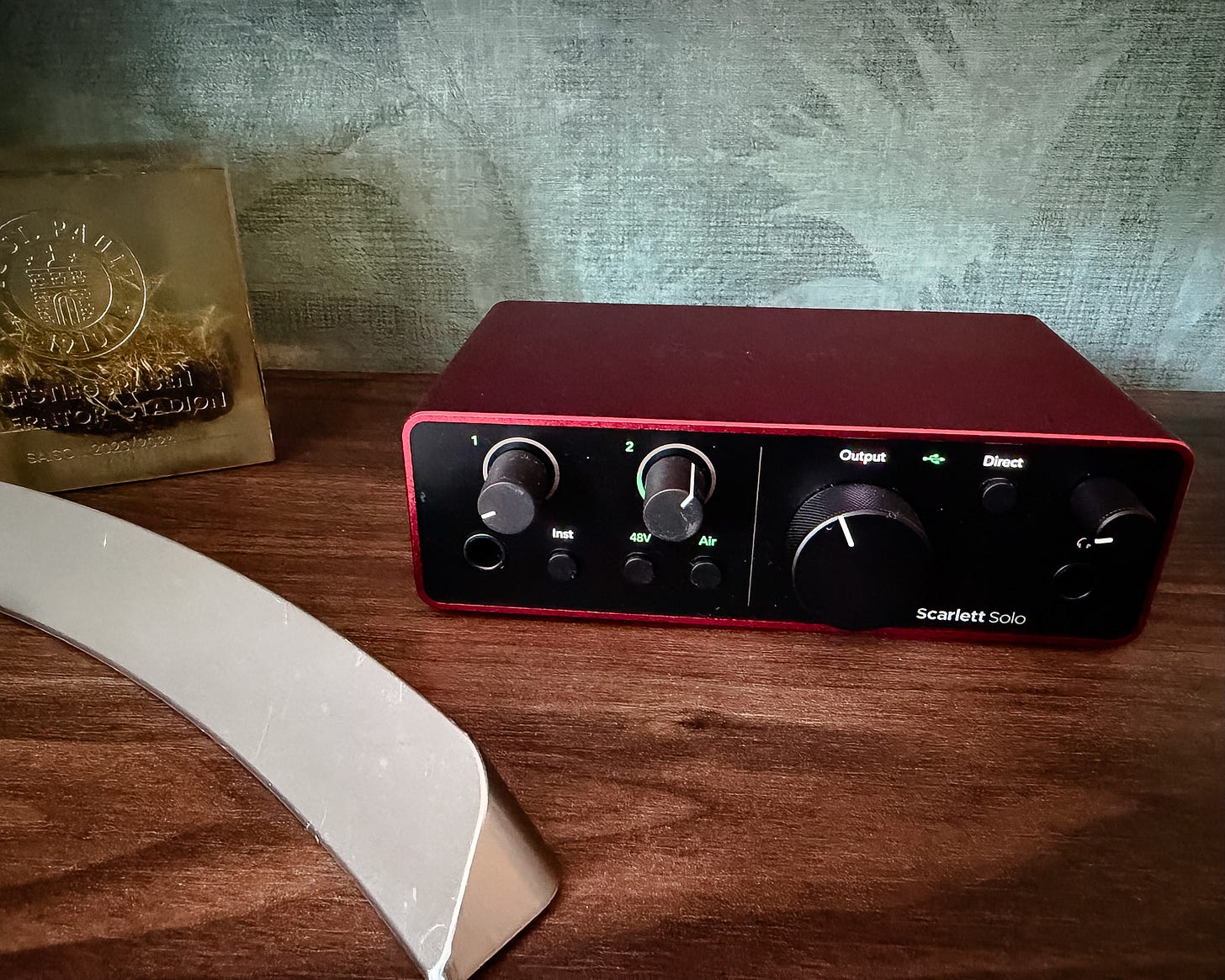 Ein rotes Focusrite Scarlett Solo USB-Audiointerface steht auf einem Holztisch. Das schwarze Frontpanel zeigt zwei Gain-Regler, Buttons für Inst, 48V und Air, einen großen Output-Regler sowie einen Kopfhöreranschluss. Der Kanal-2-Regler leuchtet grün. Links im Bild ist ein Würfel mit dem Emblem des FC St. Pauli zu sehen, im Vordergrund liegt der silberne Bügel eines Monitors. Der Hintergrund ist eine blaugrau strukturierte Wand.