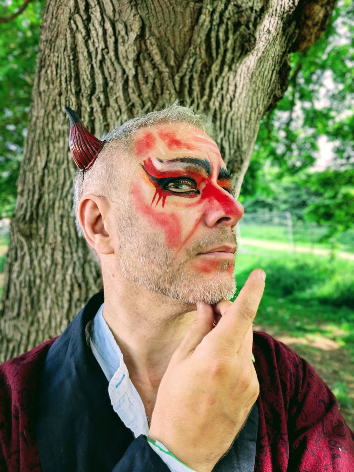 Finbarre poses beneath a large tree wearing dramatic red and black face paint, a small devil horn and a burgundy costume, one finger held thoughtfully beneath his chin. Finbarre poses beneath a large tree wearing dramatic red and black face paint, a small devil horn and a burgundy costume, one finger held thoughtfully beneath his chin.