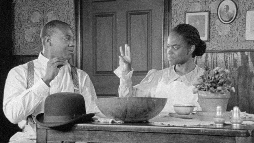 A Black Deaf couple are seated at a table at home. The woman signs 6.