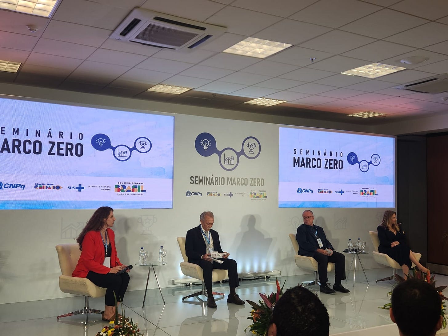 Descrição da imagem: Fotografia de um palco, onde aparecem quatro pessoas sentadas em cadeiras individuais, uma ao lado da outra, sendo duas mulheres nas pontas e dois homens ao centro. Ao fundo, há um painel onde é possível ler Seminário Marco Zero, acompanhado da logomarca do evento, constituída por 3 círculos azuis interligados, em formato de V. No primeiro, à esquerda, preenchido na cor azul, aparece uma lâmpada desenhada em branco. No círculo central, de fundo branco, aparece o desenho de um gráfico de seta evolutiva, na cor azul. No círculo que está mais à direita da imagem, também de fundo branco, há um troféu com uma estrela inscrita, ambos na cor azul.
