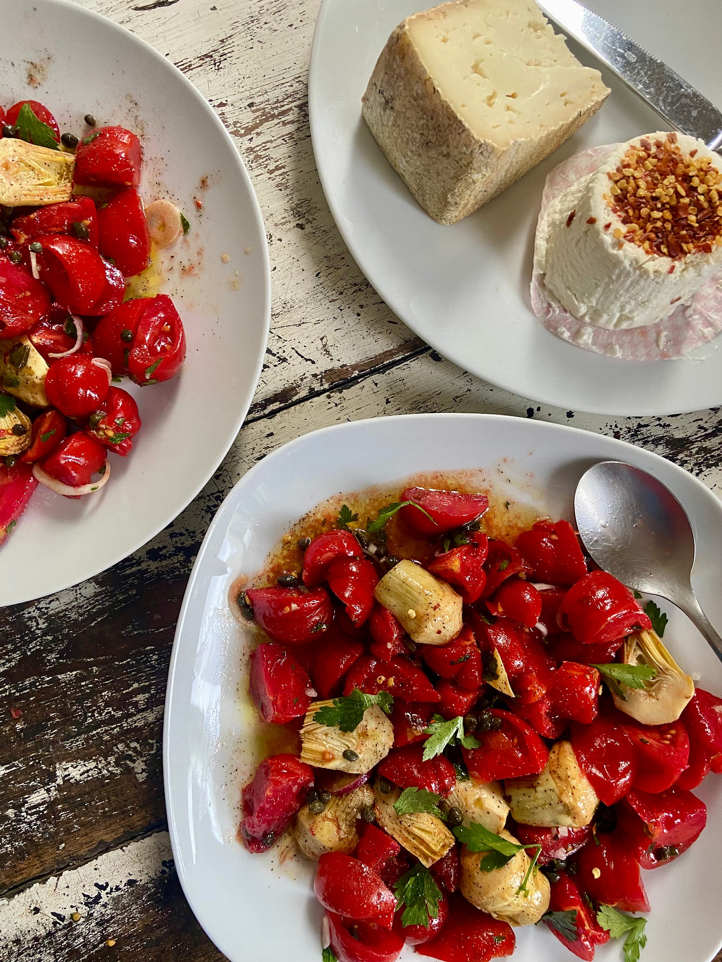 The image shows two bowls filled with oily tomatoes parsley and artichoke hearts. A big piece of cheese sits in the top right cornner, covered in chili flakes. 