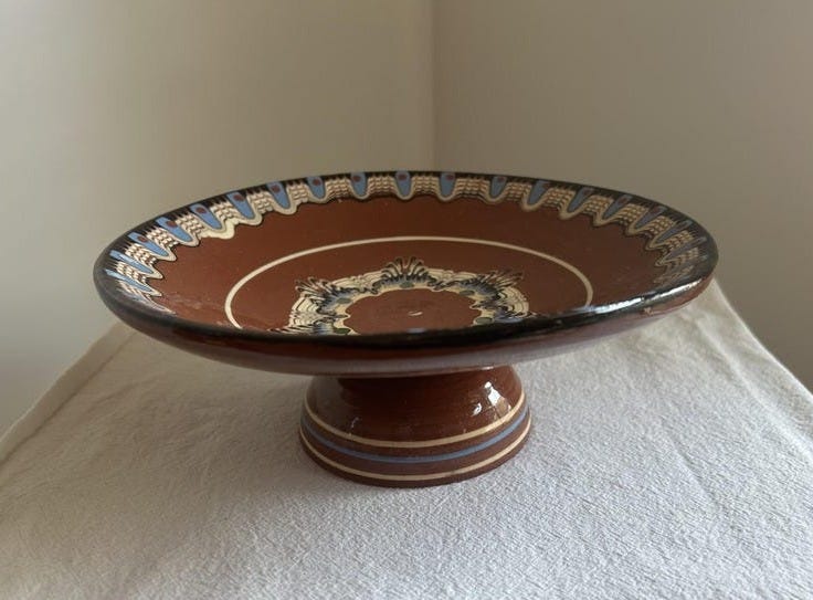 May include: A brown ceramic pedestal bowl featuring a wide, shallow basin and a sturdy base. The rim is decorated with a repeating blue, white, and black pattern, while the center of the bowl displays a detailed floral-inspired motif in matching colors.