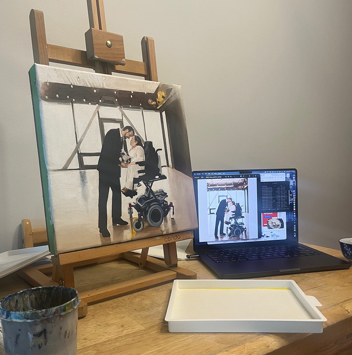 In an echo of the studio layout, the dining table has the wedding painting on the same desktop easel and an almost identical state of incompleteness. Beside it sits the laptop with the reference photo of the bride and groom. On the table is a waiting blank paint palette and a paint covered water container. In an echo of the studio layout, the dining table has the wedding painting on the same desktop easel and an almost identical state of incompleteness. Beside it sits the laptop with the reference photo of the bride and groom. On the table is a waiting blank paint palette and a paint covered water container.
