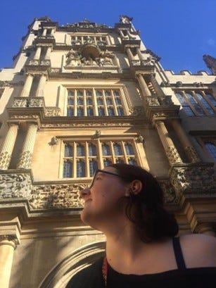 A photograph shows a brunette woman with glasses wearing an off-the-shoulder black top, looking upwards at a grand, tall building of the University of Oxford.