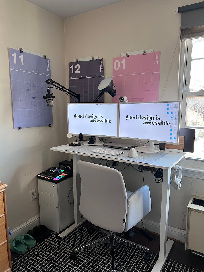 This is a photo of a clean and well-organized home office setup. The workspace includes:  A standing desk with a white surface, featuring dual monitors displaying the message "good design is accessible." A white ergonomic chair on wheels. Mounted calendars on the wall in a gradient color scheme for November, December, and January. An adjustable microphone arm, suggesting the space may be used for recording or streaming. Desk accessories like a lamp, small gadgets, and a keyboard/mouse setup. A small drawer unit to the left of the desk, topped with what appears to be a soundboard or control panel with colorful buttons. A patterned rug under the desk area, adding texture to the setup. A window to the right, providing natural light.