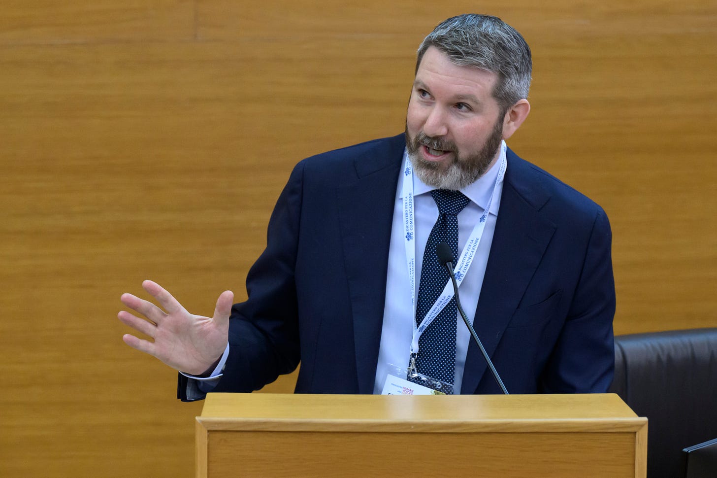 A bearded man in a dark navy suit and dotted tie speaks at a light wood podium, gesturing with one hand. He is wearing a conference lanyard reading "Dicastero per la Comunicazione." A microphone is mounted on the podium in front of him.