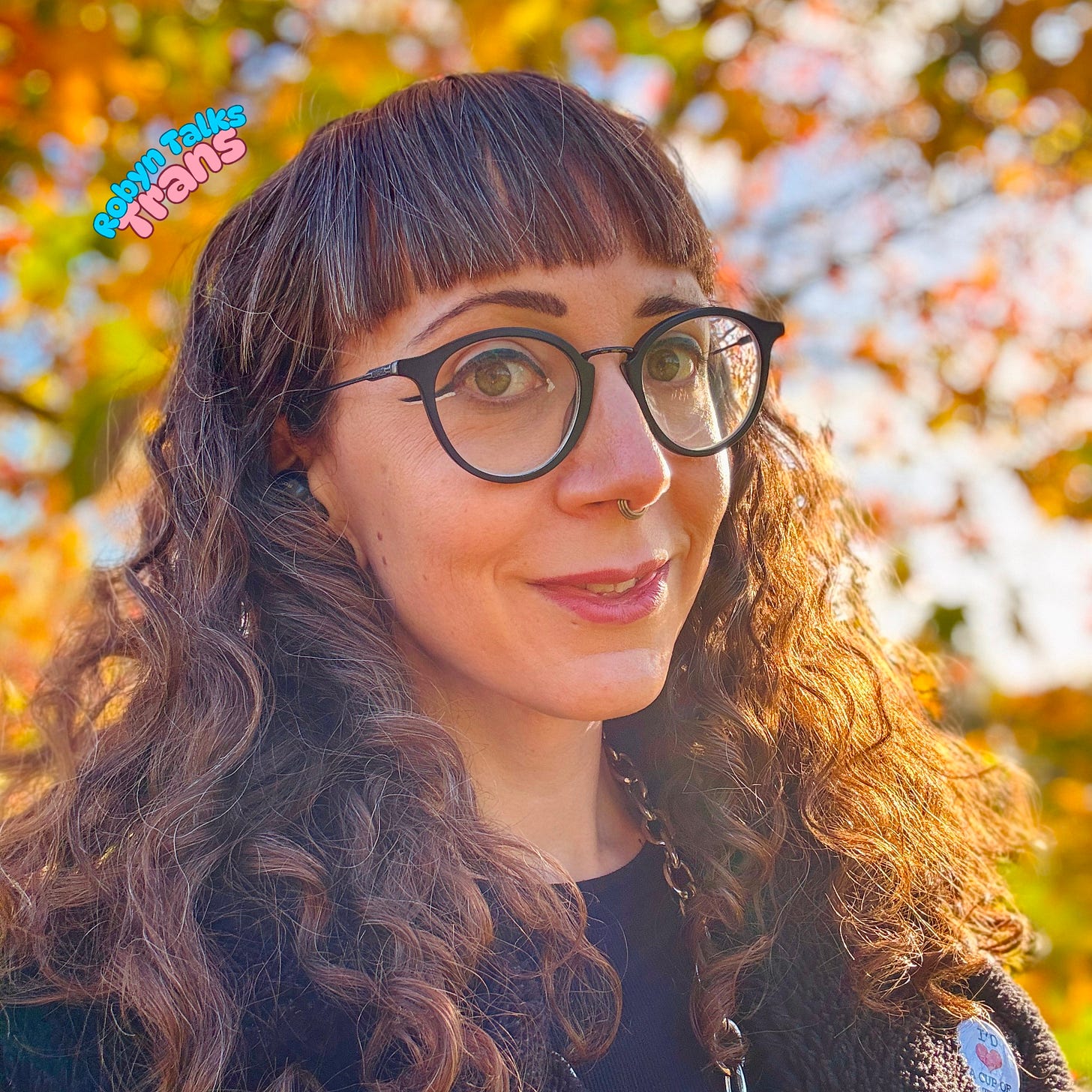 A photo of Robyn with orange and brown leaves in the background, and the sun shining. She is smiling and looking at the camera A photo of Robyn with orange and brown leaves in the background, and the sun shining. She is smiling and looking at the camera
