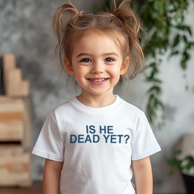 Young toddler girl with blonde hair in pigtails smiles directly at the camera with wide eyes and a big grin. She wears a plain white t-shirt featuring the text Is He Dead Yet? in large blue capital letters across the chest. The background includes a textured gray wall, green leafy plants, and wooden furniture elements.