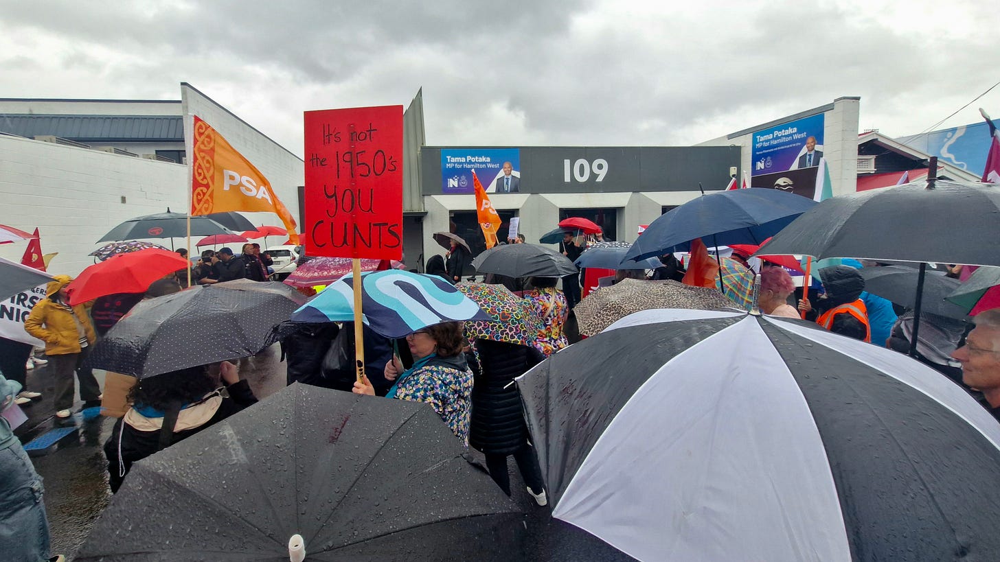 image of people holding umbrellas outside a local mp's office. one sign reads, its not the 1950s you cunts