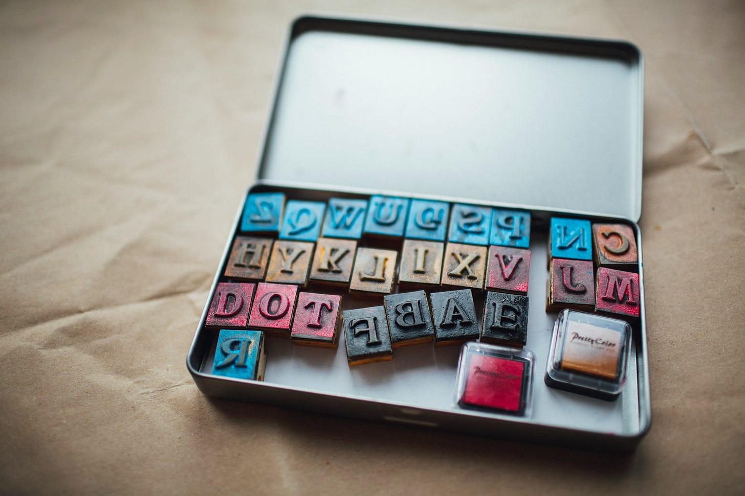 Una scatola di latta aperta contenente una serie di caratteri mobili in legno con lettere dell'alfabeto. I caratteri mobili hanno la base in legno e la parte superiore colorata di blu, nero e rosso. Le lettere sono incise sulla parte superiore e mostrano anche residui di inchiostro colorato. Le lettere sono disposte in modo casuale, non in ordine alfabetico. Accanto ai caratteri mobili ci sono due piccoli tamponi d'inchiostro, uno rosso e uno nero. Una scatola di latta aperta contenente una serie di caratteri mobili in legno con lettere dell'alfabeto. I caratteri mobili hanno la base in legno e la parte superiore colorata di blu, nero e rosso. Le lettere sono incise sulla parte superiore e mostrano anche residui di inchiostro colorato. Le lettere sono disposte in modo casuale, non in ordine alfabetico. Accanto ai caratteri mobili ci sono due piccoli tamponi d'inchiostro, uno rosso e uno nero.