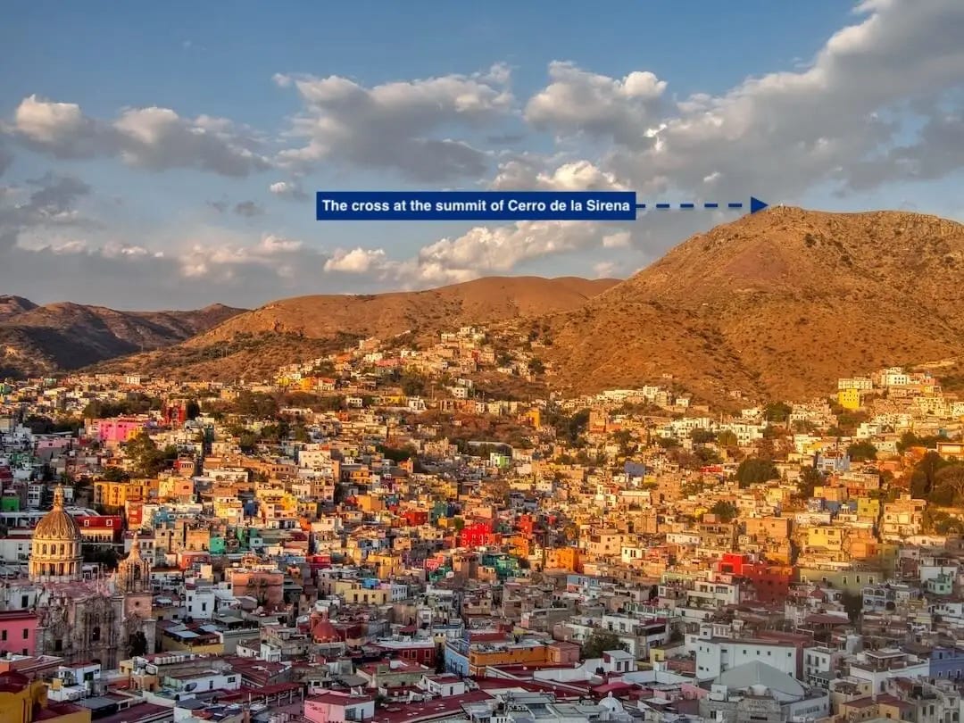Sun shines on a hillside with a white cross at its summit above a town of coloured buildings