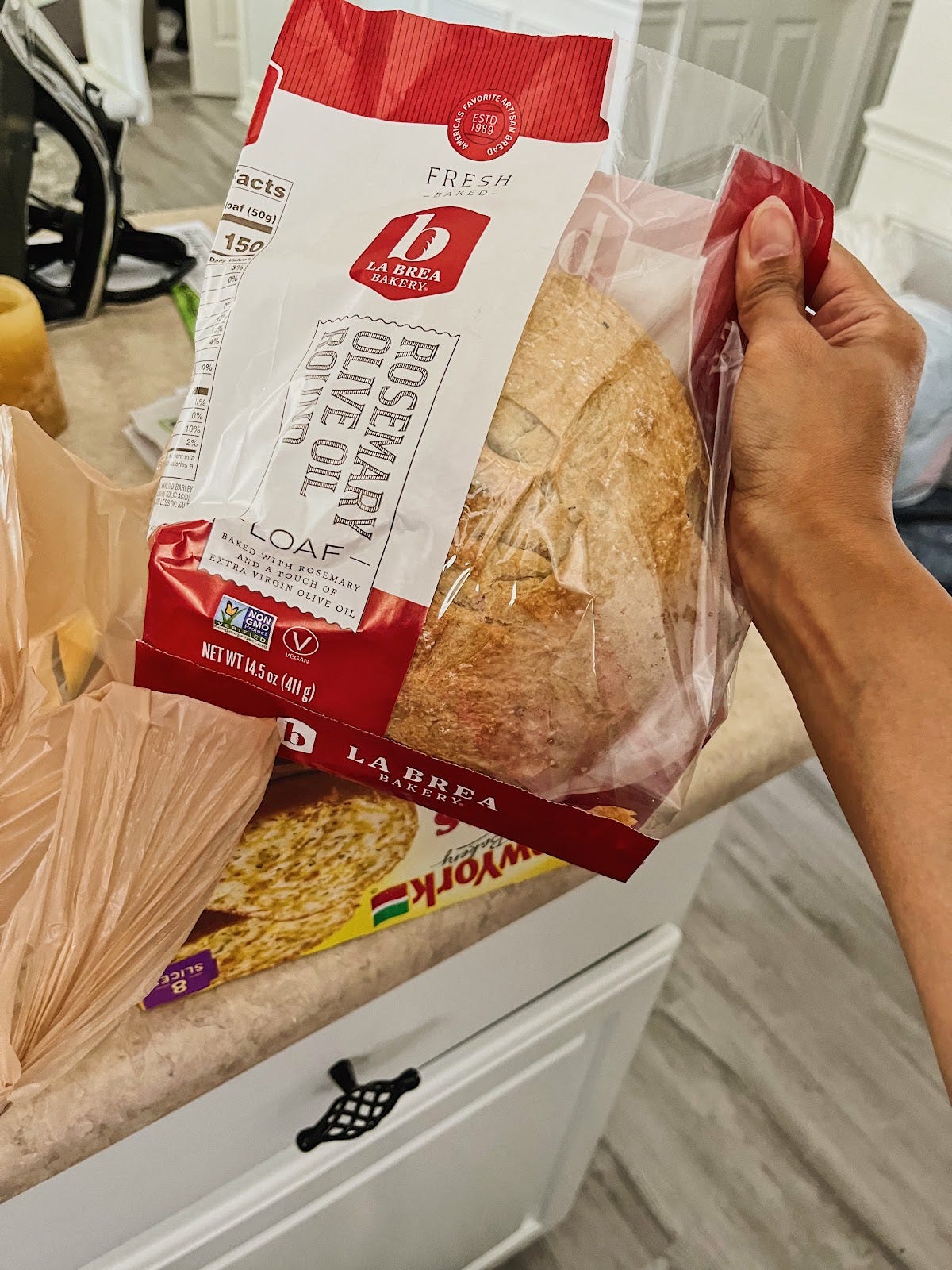 A hand holding a loaf of rosemary olive oil bread in a bag with grocery bags behind on a kitchen counter