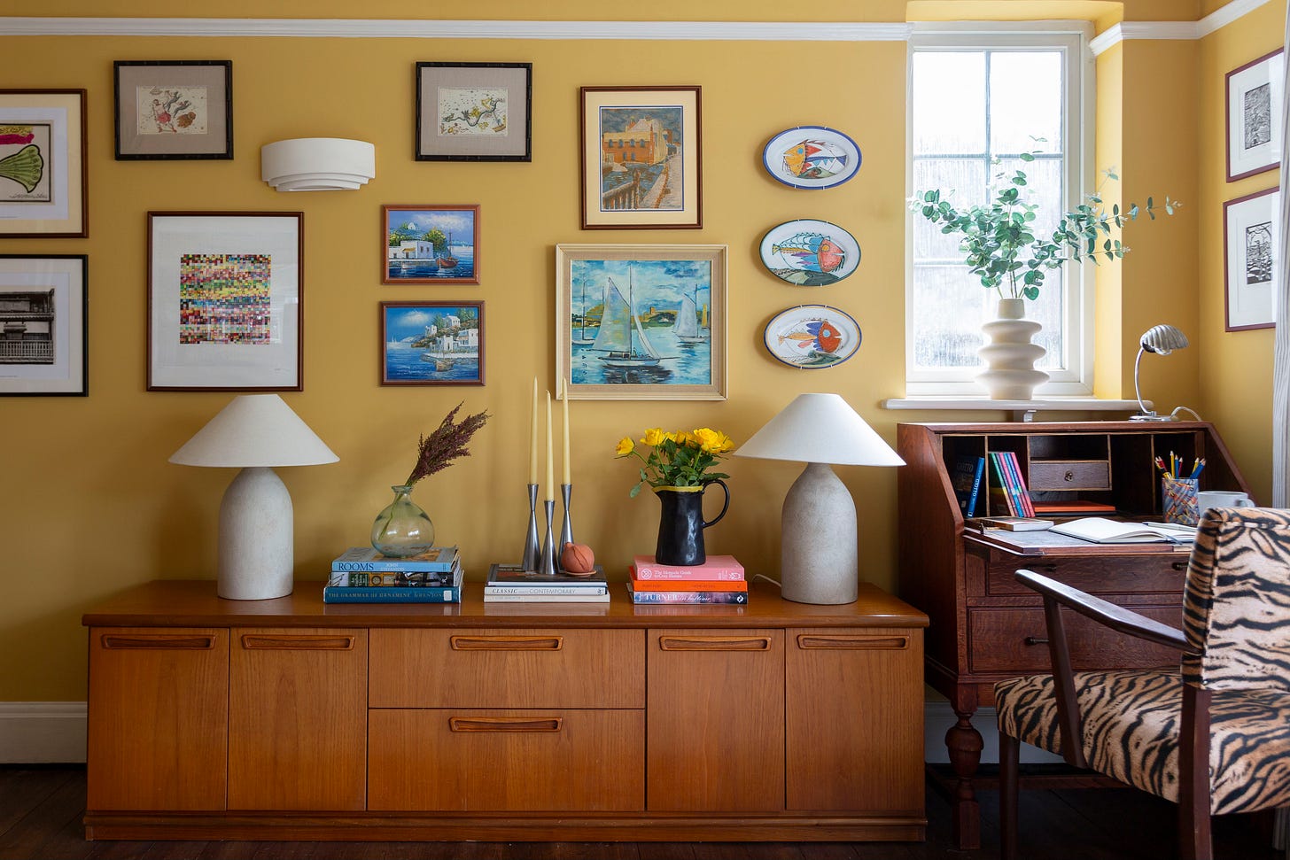 Yellow living room with gallery wall, sideboard and bureau. Two lamps and books on sideboard