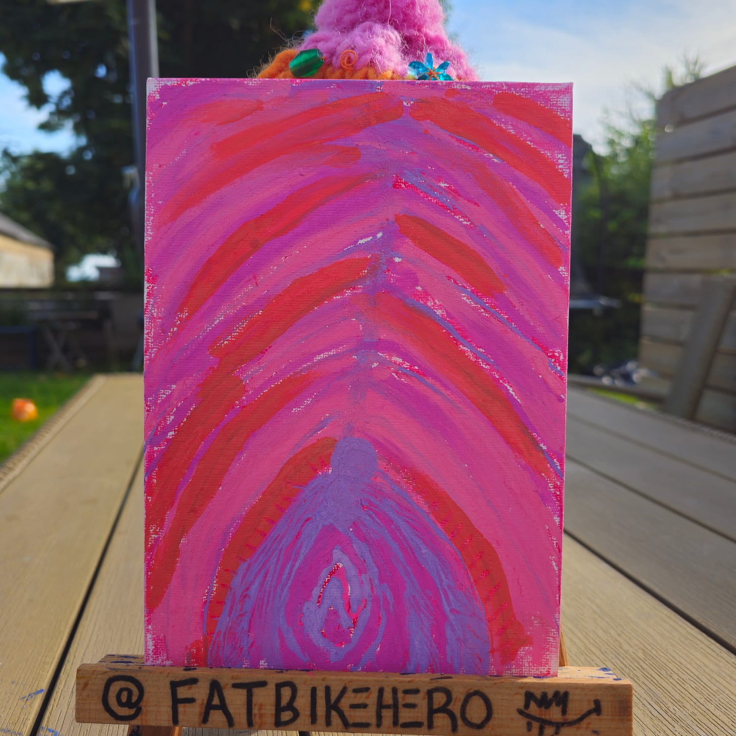 Alt Text: A vertical painting by Fatbike Hero featuring arching bands of pink, red, and orange paint converging at a central spine. At the bottom, a purple, loop-like shape resembles a flower bud or anatomical diagram. The painting rests on a wooden easel signed "@FATBIKEHERO". Alt Text: A vertical painting by Fatbike Hero featuring arching bands of pink, red, and orange paint converging at a central spine. At the bottom, a purple, loop-like shape resembles a flower bud or anatomical diagram. The painting rests on a wooden easel signed "@FATBIKEHERO".