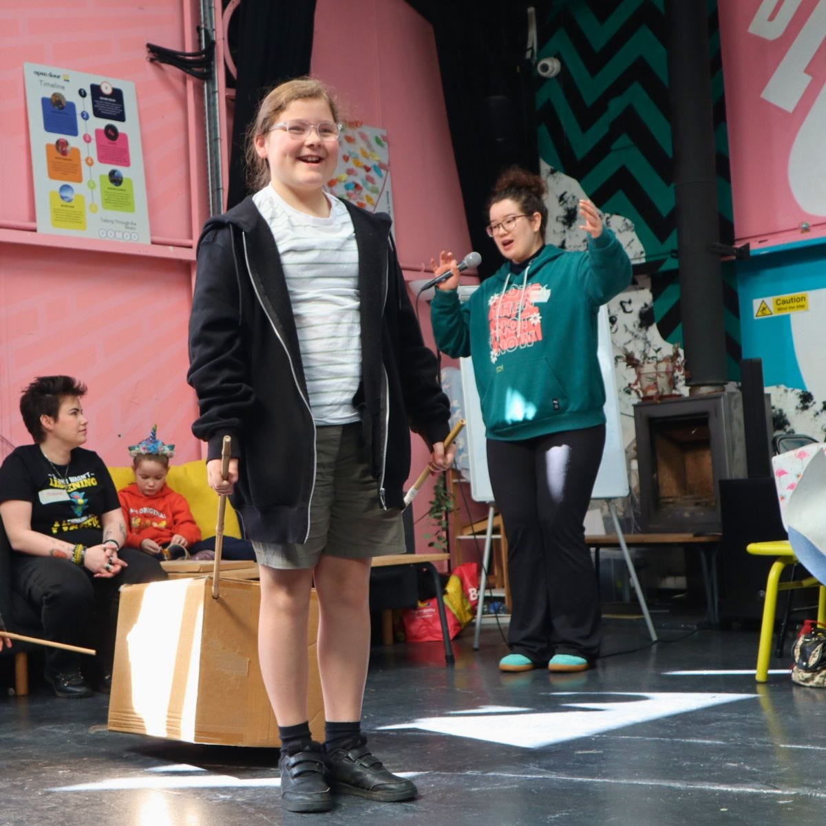 A child holds two bamboo sticks and a box behind them, while Mai-Leen speaks on the microphone. The child is smiling
