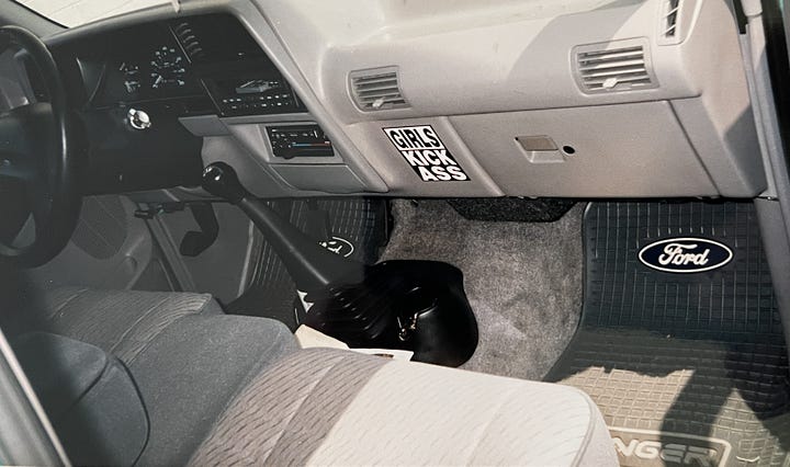 old blue Ford pickup truck with white grill, and gray interior of 1993 truck cab with "Girls Kick Ass" sticker on dash