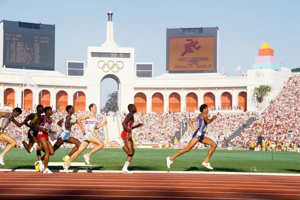 Competitors on a running track, with the peristyle end of the Los Angeles Memorial Coliseum in the background. Scoreboards are on either side of an archway, with the Olympic rings above it.