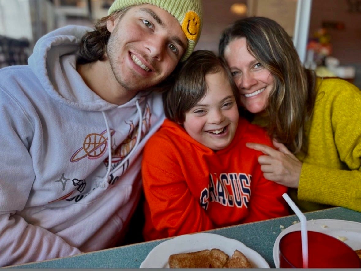 Three white people with their heads together, smiling. On the left, a young man wearing a neon yellow beanie; in the middle, a boy with Down syndrome, wearing an orange Syracuse sweatshirt'; on the right, a middle aged woman with long brown hair, wearing a yellow sweater. Three white people with their heads together, smiling. On the left, a young man wearing a neon yellow beanie; in the middle, a boy with Down syndrome, wearing an orange Syracuse sweatshirt'; on the right, a middle aged woman with long brown hair, wearing a yellow sweater.