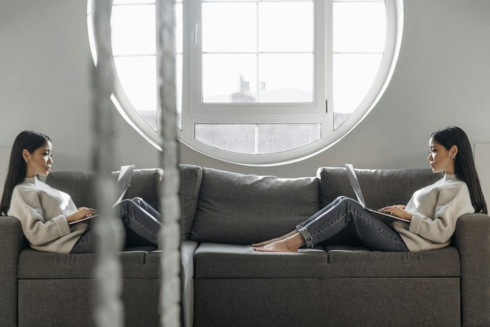 A woman sitting on a couch with a laptop, with a mirror image of herself on the other end of the couch