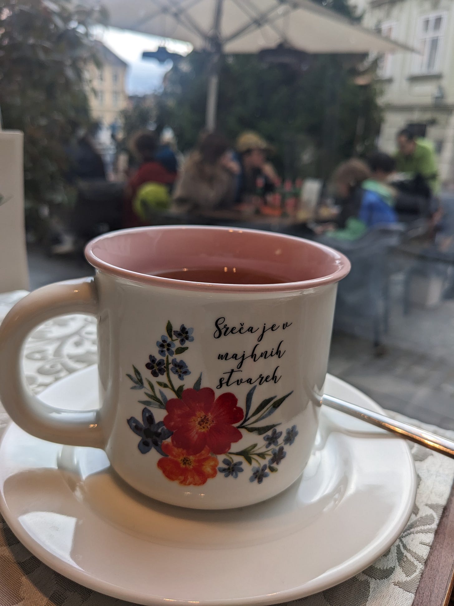  A white cup filled with tea has flowers on it and a message that reads: “Happiness is in small things” in Slovenian. The cup is sitting on a table by the café window.