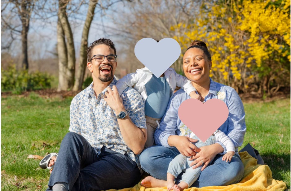 A bright outdoor family portrait in a backyard. Two adults sit on a yellow blanket on the grass, smiling widely at the camera. Mariella's husband, on the left, wears glasses, a light blue floral short-sleeve shirt, dark pants, and a watch. Mariella, on the right, wears a blue striped blouse with floral embroidery and jeans. Two small children are with them: one child stands between the adults with their face covered by a blue heart shape, and another child sits on the woman’s lap with their face covered by a pink heart shape. Behind them are bare trees, green grass, and a large bush with bright yellow flowers, giving the image a warm springtime feel.