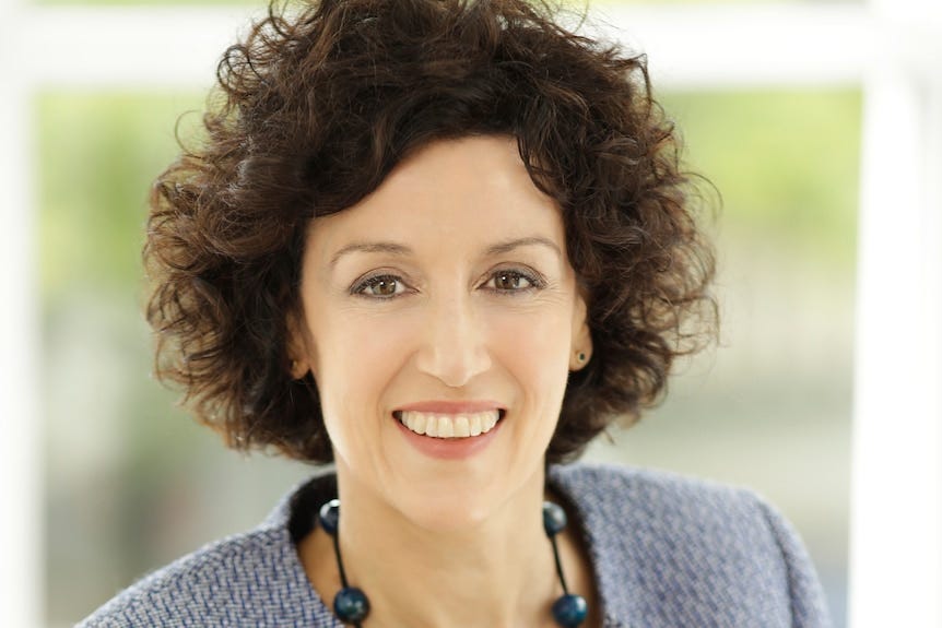A professional headshot of a woman with brown curly hair and a suit jacket smiling at the camera A professional headshot of a woman with brown curly hair and a suit jacket smiling at the camera