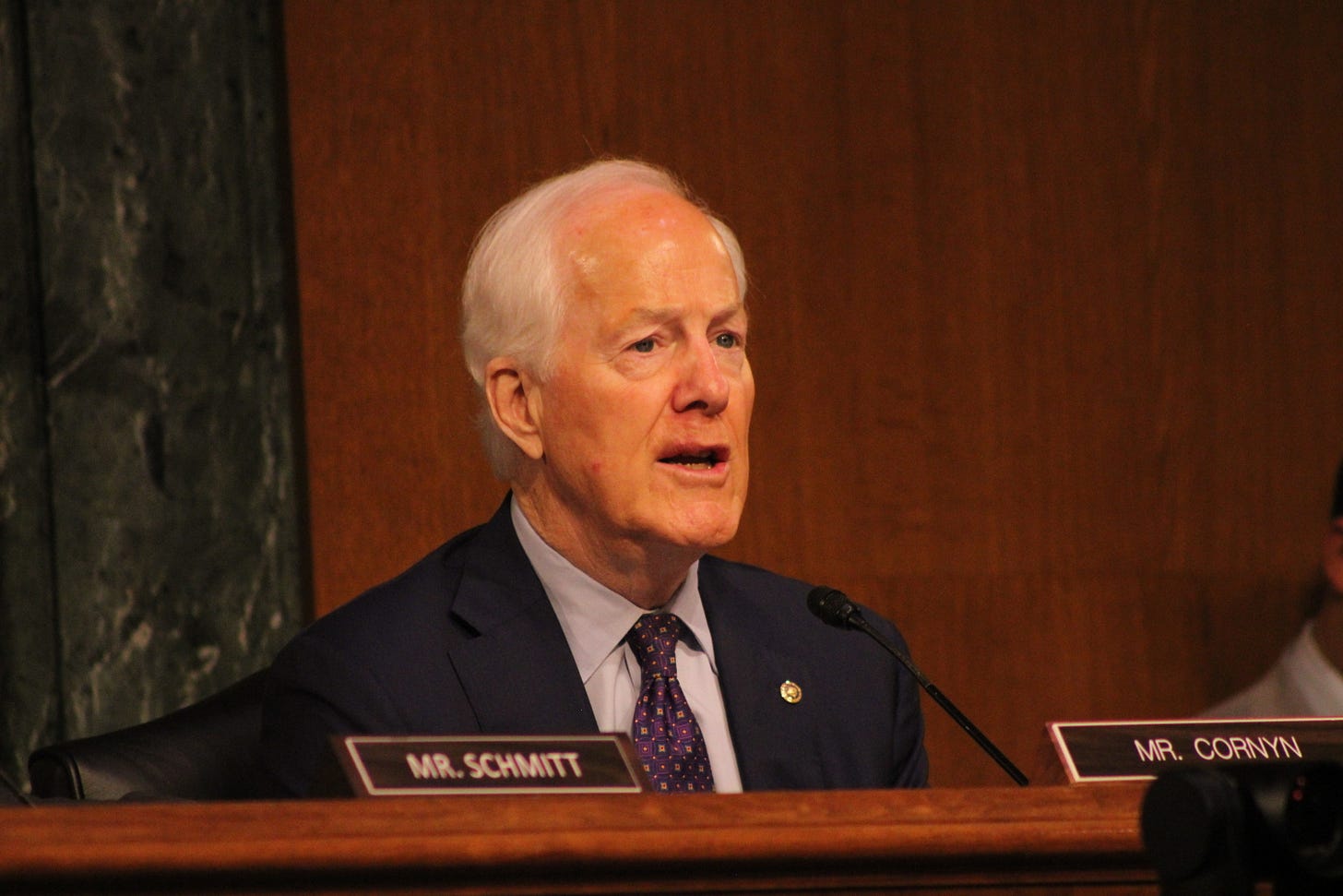 A white man in a suit asks a question from a wooden dais. Photo: Logan Johnson © www.askapol.com A white man in a suit asks a question from a wooden dais. Photo: Logan Johnson © www.askapol.com