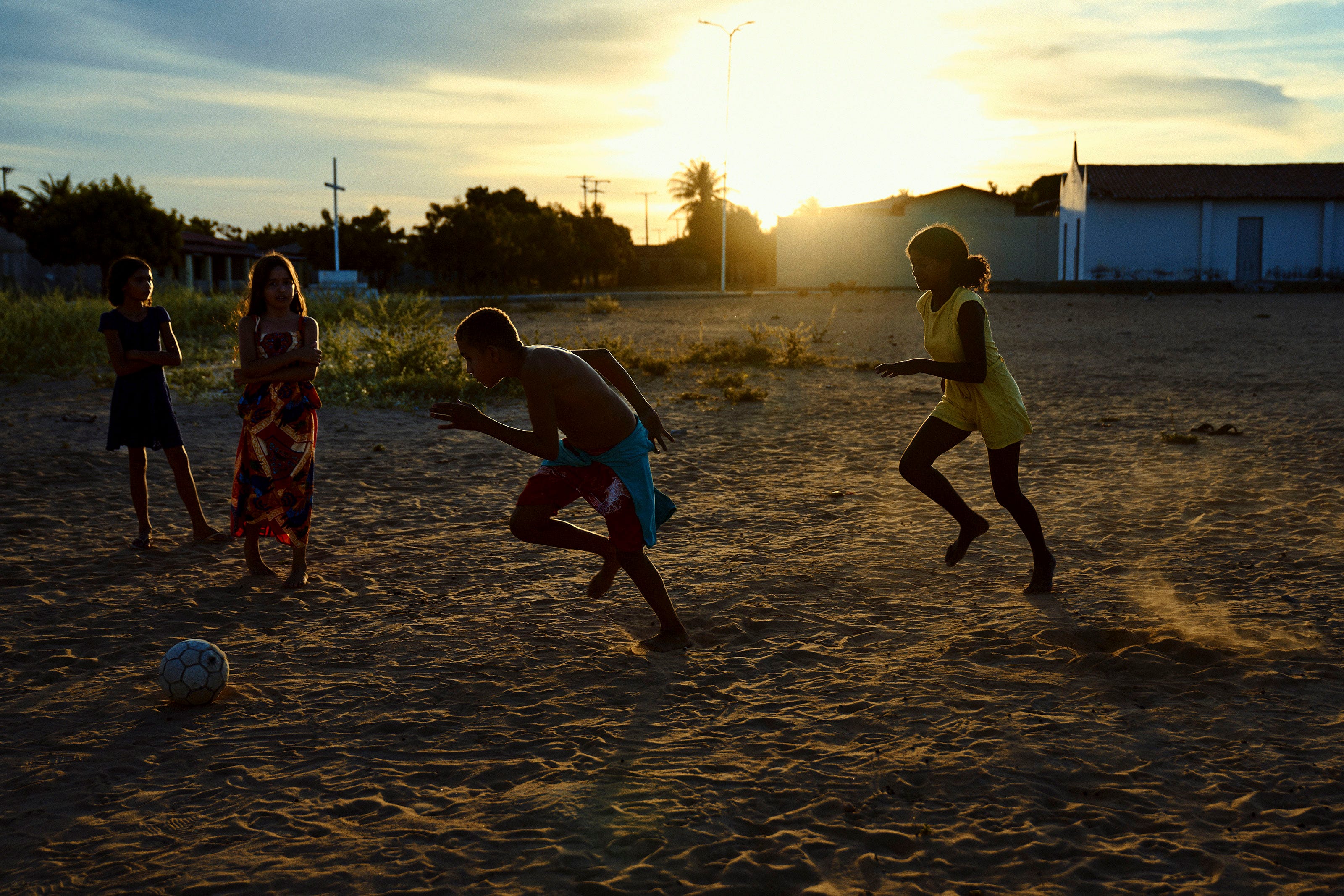 Four children play soccer on a sandy field at sunset, with two chasing the ball while two others watch nearby. The warm sunlight creates long shadows and silhouettes against a rural backdrop.