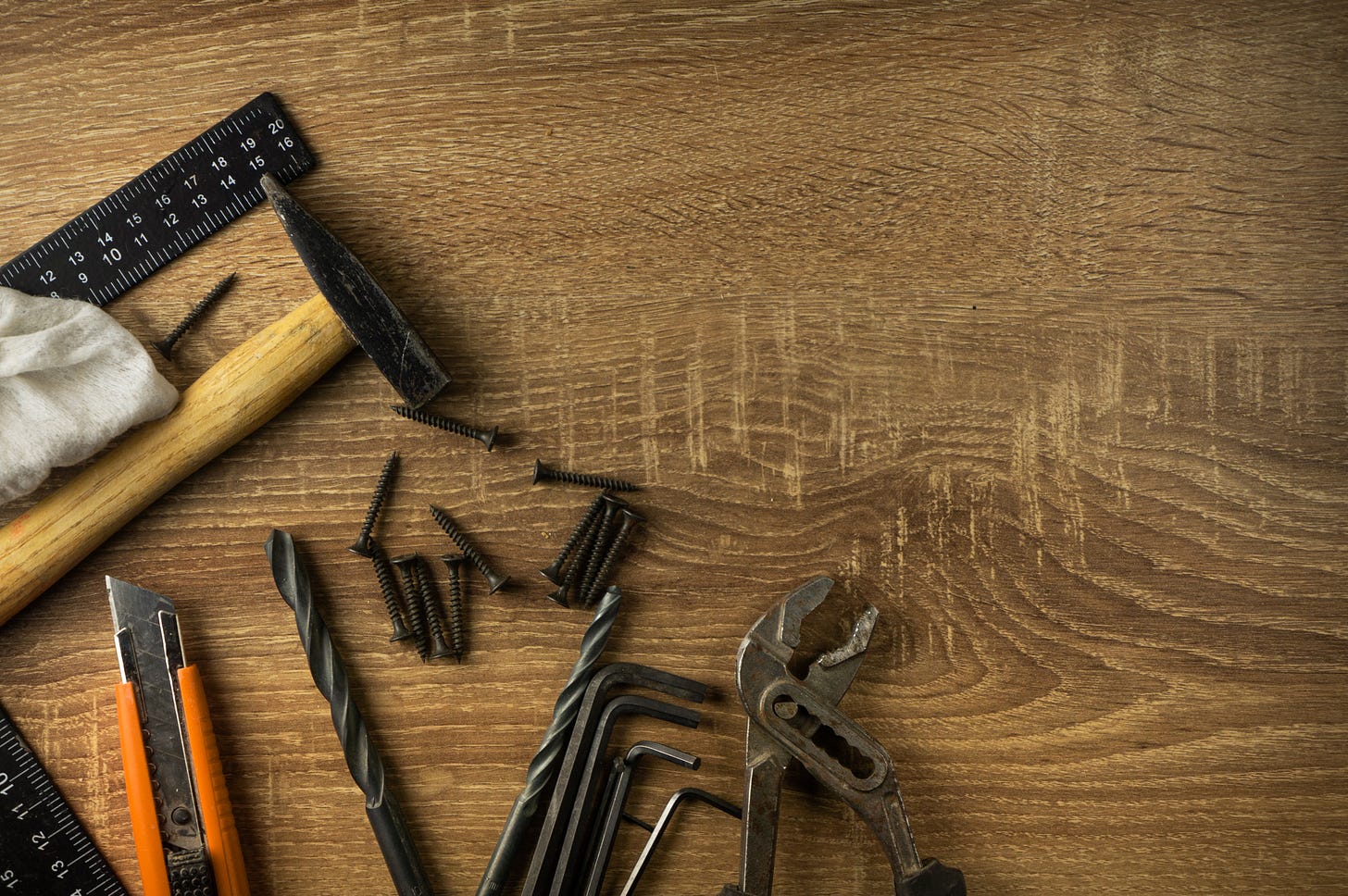 A number of hand tools rest on a wooden table - ruler, nails, drill bit, pliers, blade, hammer.