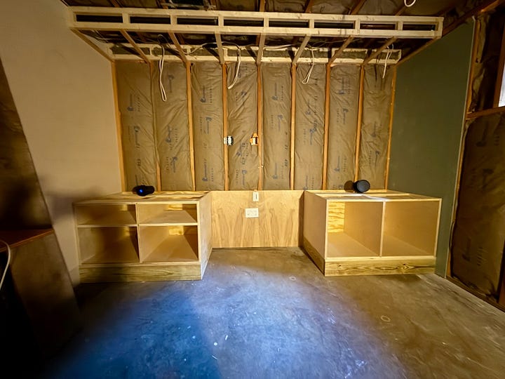 Two photos of basement cabinetry construction. The first shows a close-up of a plywood cabinet box with tools on top and storage bins on the floor. The second shows a wider view with two cabinet bases installed against an insulated wall, wiring exposed overhead.