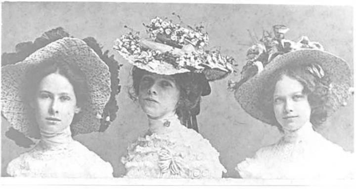 Black and white antique photo of three girls dressed up in fancy dress with elaborate, dressy hats worn in the 1800s.