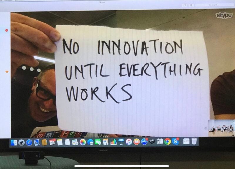 This image shows a video call where a man is holding up a handwritten paper sign. The sign says in bold black letters  "NO INNOVATION UNTIL EVERYTHING WORKS