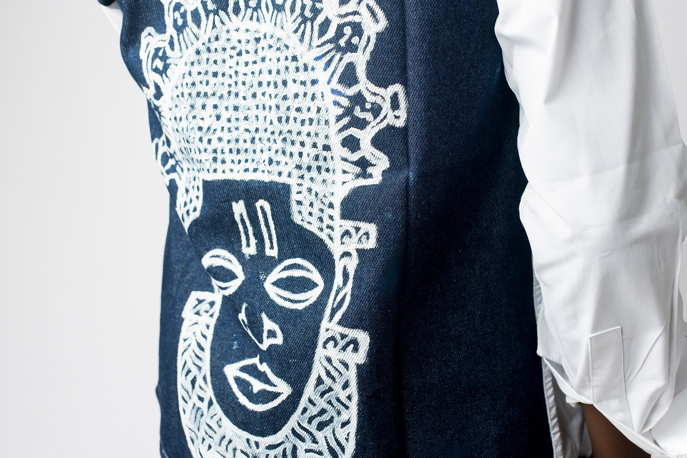 A hand-painted ancient African mask motif in white across the indigo back panel of a Heritage Collection garment, photographed in the studio A hand-painted ancient African mask motif in white across the indigo back panel of a Heritage Collection garment, photographed in the studio